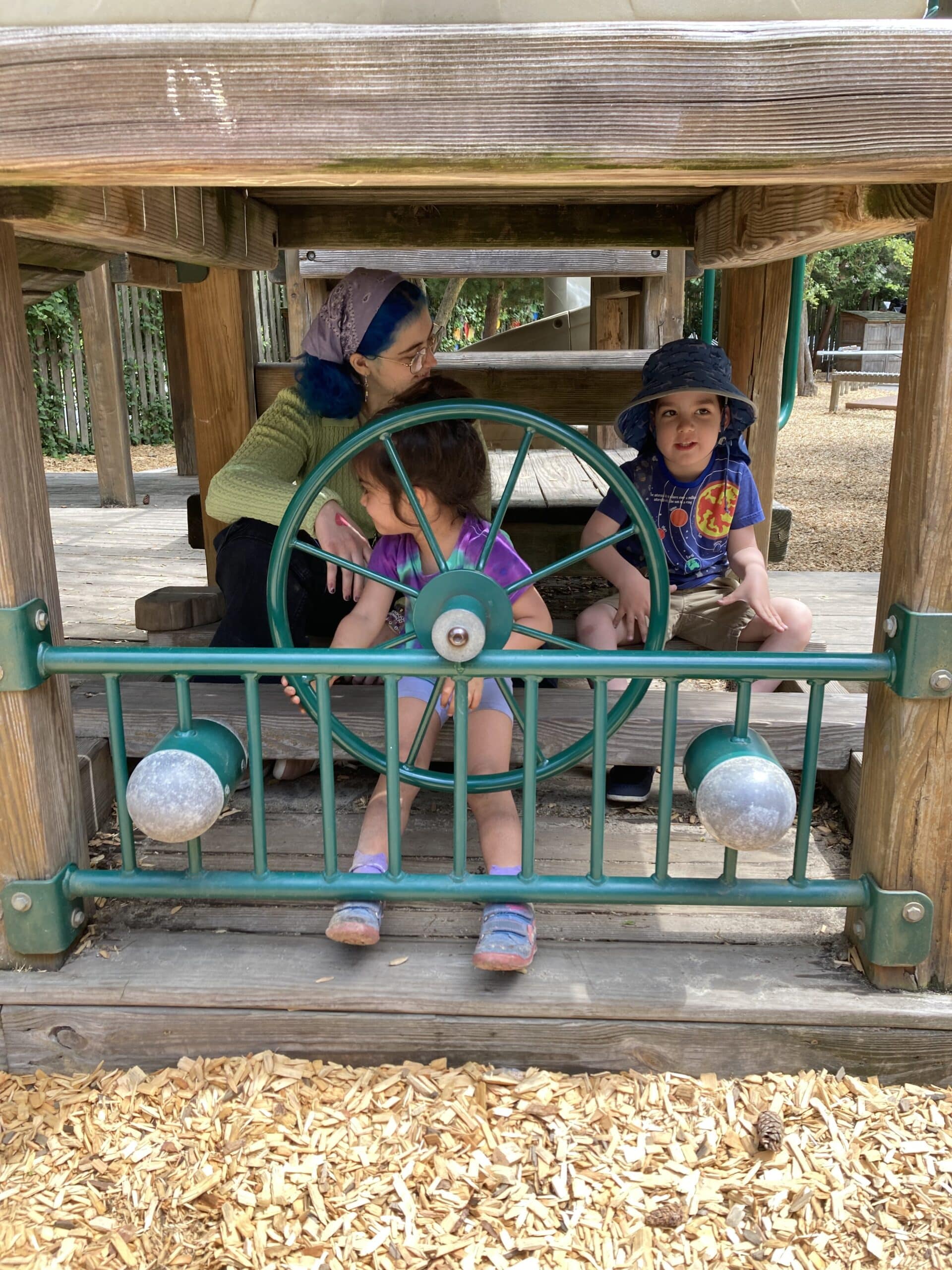 Two campers and a counsellor sit in a life size wooden train in the playground