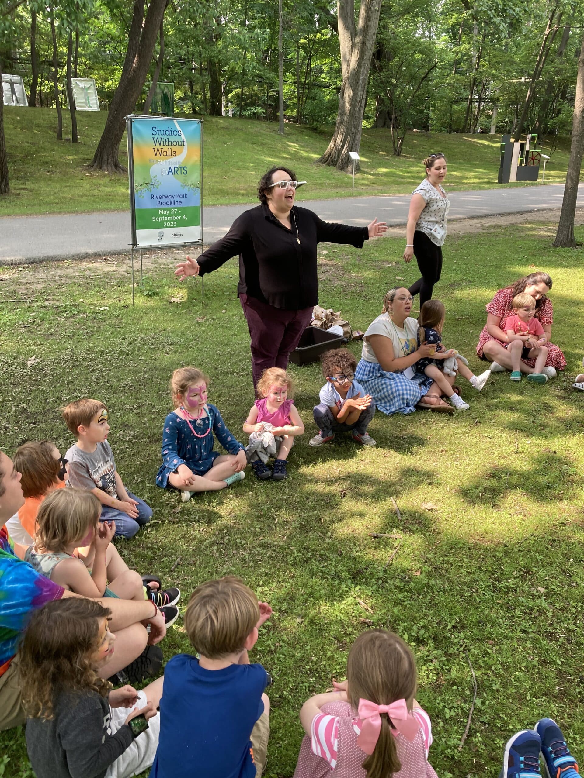 Rabbi Suzie Jacobson standing with arms outstretched beside a group of children sitting in the park