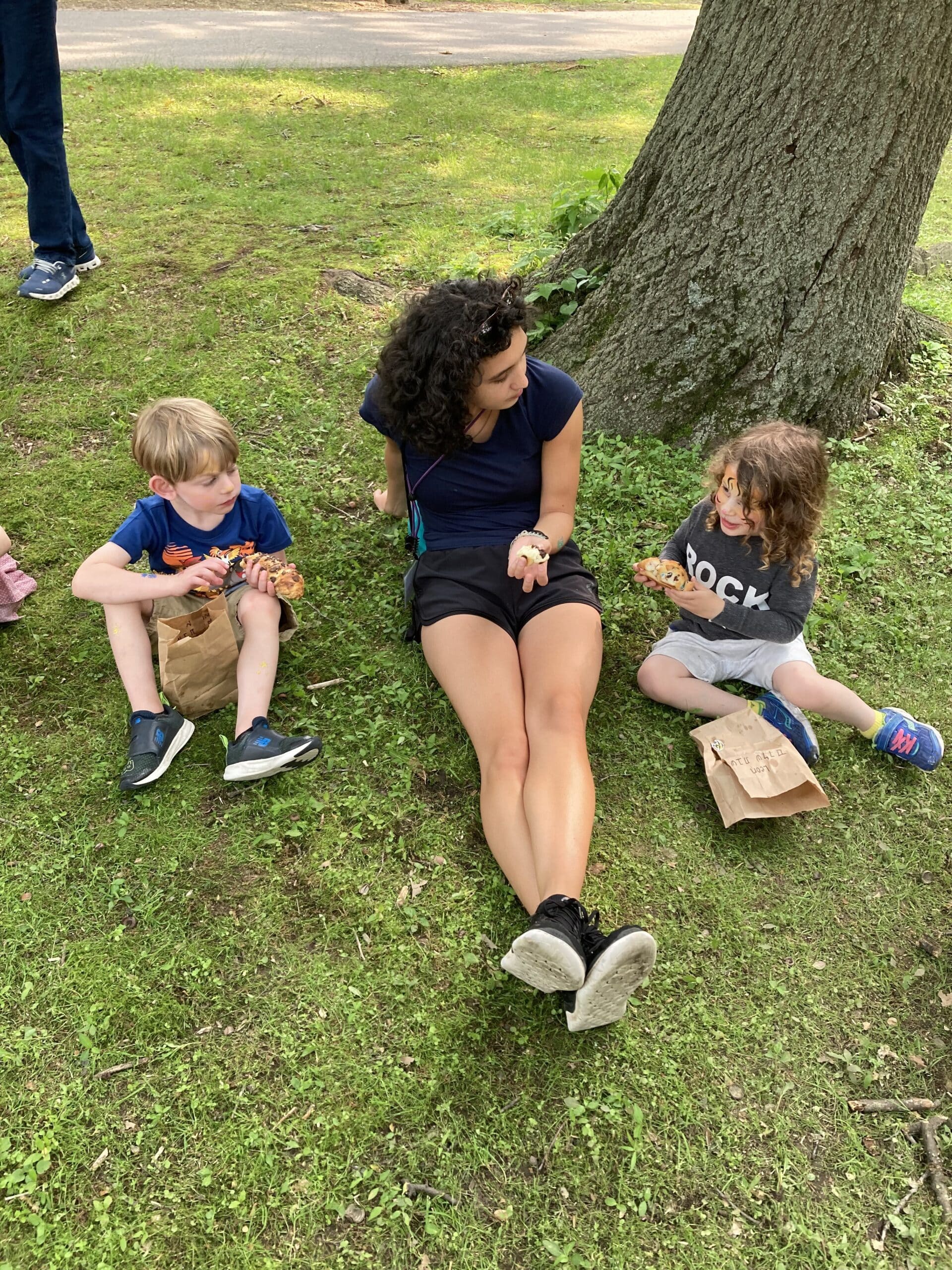 A camp counsellor and two kids eating lunch in the park beside a big tree