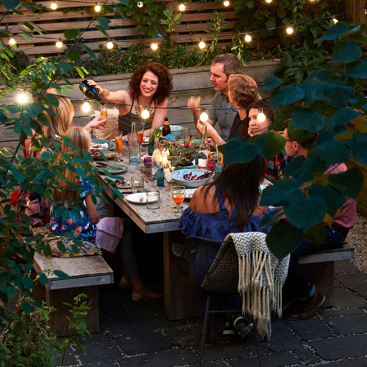 A group of people in their 40s eatingat an outdoor table with string lights and green plants